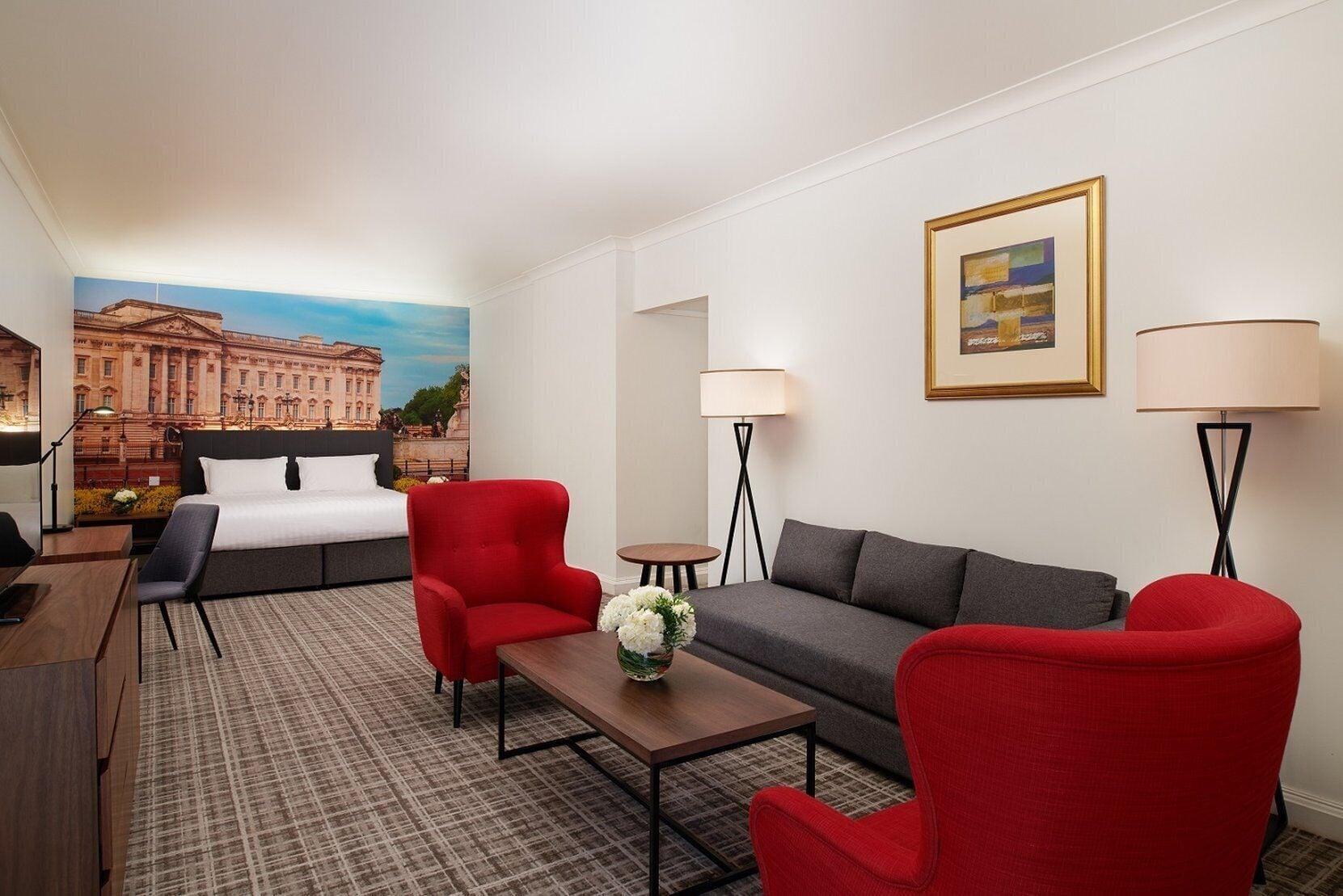 Millennium Hotel And Conference Centre Gloucester London Exterior photo The photo shows a modern hotel room featuring a cozy seating area. There are two bright red armchairs and a gray sofa arranged around a wooden coffee table, which has a small vase of flowers on it. Behind the seating area, there is a large bed with a
